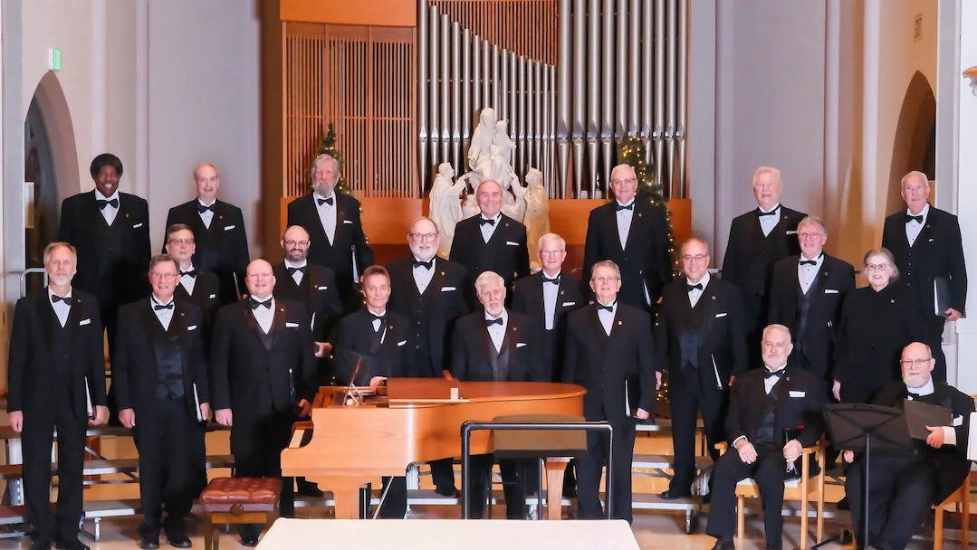 Members of the Chiaroscuro Men's Chorus lined up on risers in front of piano for concert performance